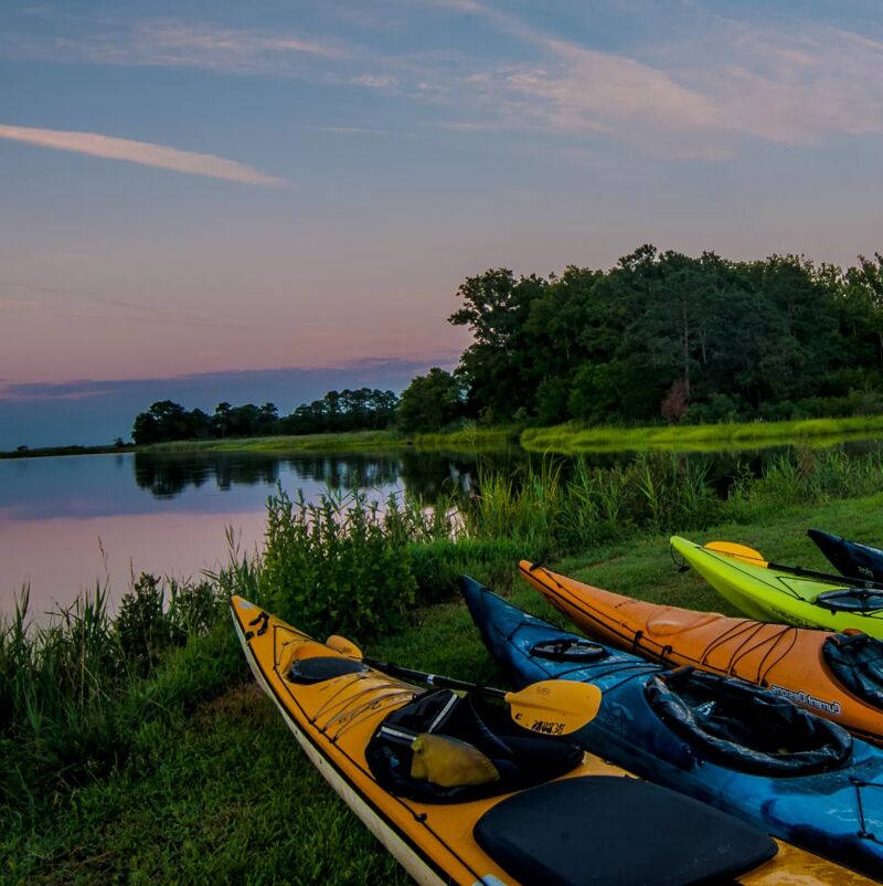 The image shows several kayaks resting on a grassy bank next to a calm body of water. The kayaks are various colors, including yellow, orange, and blue. In the background, there's a line of trees and a colorful sky, suggesting it might be dusk or dawn. The scene evokes a sense of tranquility and outdoor recreation.